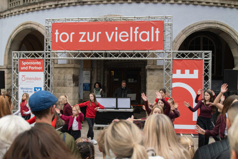 Vor einem historischen Gebäude treten mehrere Kinder und Jugendliche in roten und dunkelroten Oberteilen auf. Zahlreiche Zuschauerinnen und Zuschauer verfolgen die Darbietung. Über der Bühne hängt ein großes rotes Banner mit der Aufschrift „tor zur vielfalt“.