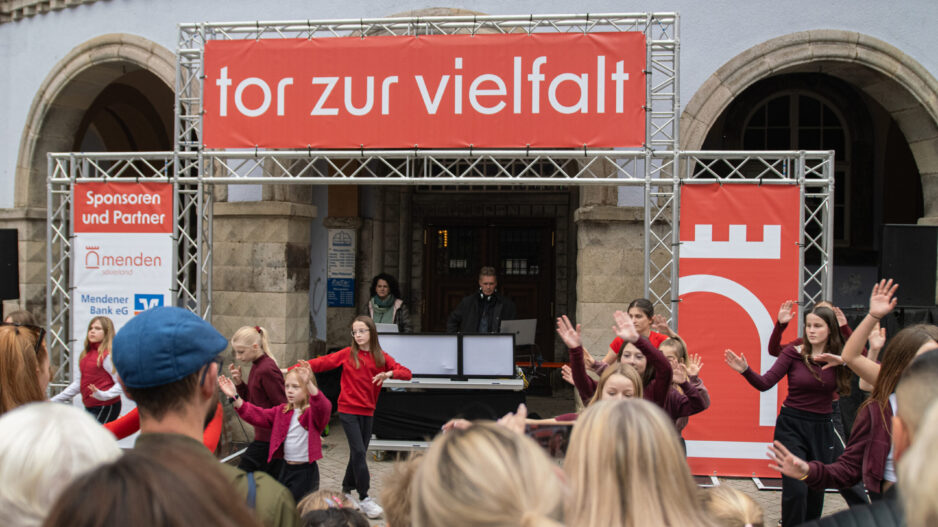 Vor einem historischen Gebäude treten mehrere Kinder und Jugendliche in roten und dunkelroten Oberteilen auf. Zahlreiche Zuschauerinnen und Zuschauer verfolgen die Darbietung. Über der Bühne hängt ein großes rotes Banner mit der Aufschrift „tor zur vielfalt“.