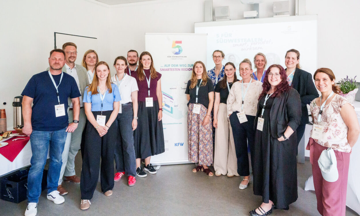 Eine Gruppe von 14 Personen steht für ein gemeinsames Foto in einem hellen Veranstaltungsraum. Die Teilnehmenden tragen Namensschilder an Schlüsselbändern. Im Hintergrund stehen ein Roll-up-Banner der Initiative „5 für Südwestfalen – Smart Cities“ und eine Präsentationsleinwand.