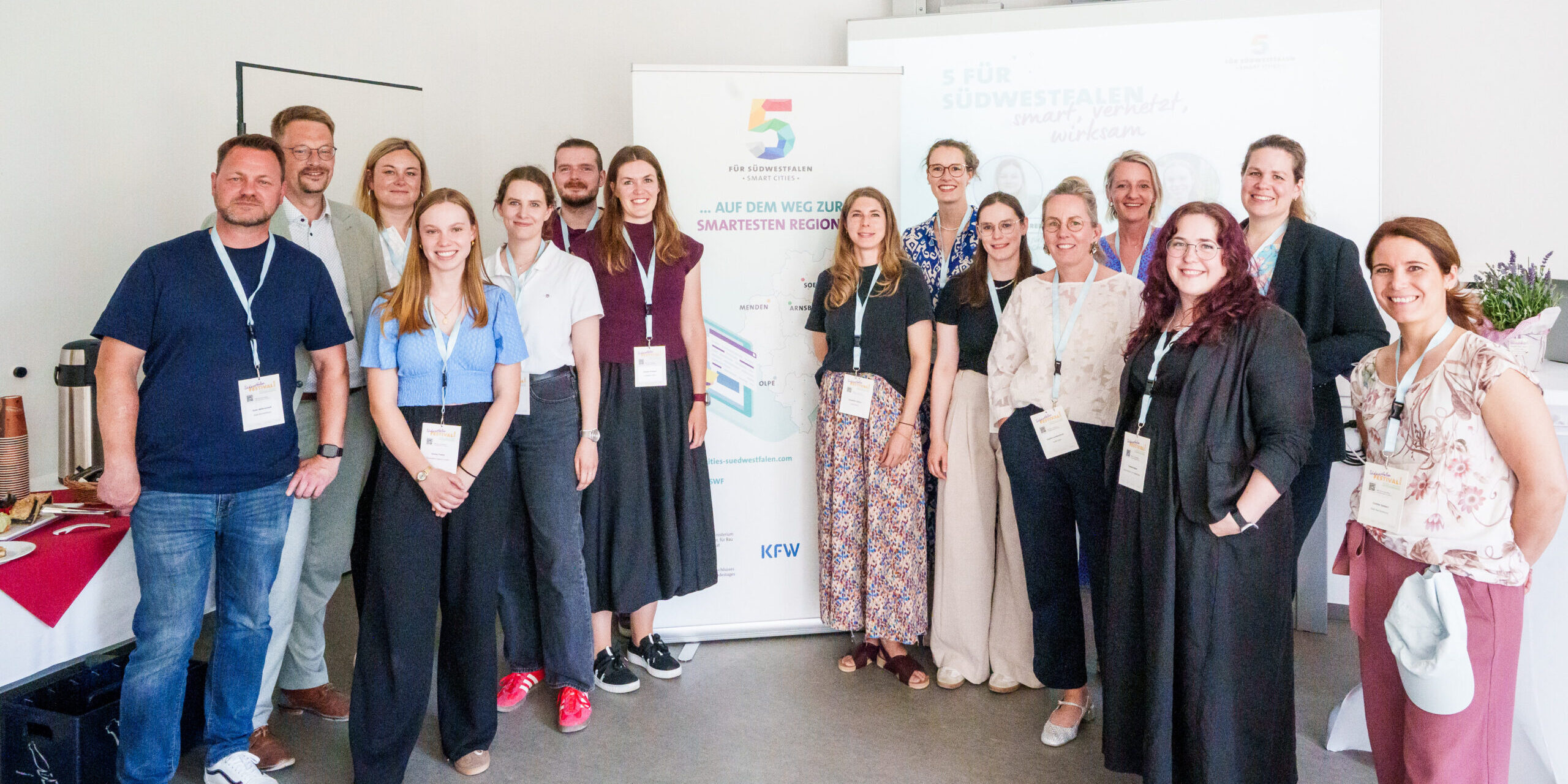 Eine Gruppe von 14 Personen steht für ein gemeinsames Foto in einem hellen Veranstaltungsraum. Die Teilnehmenden tragen Namensschilder an Schlüsselbändern. Im Hintergrund stehen ein Roll-up-Banner der Initiative „5 für Südwestfalen – Smart Cities“ und eine Präsentationsleinwand.