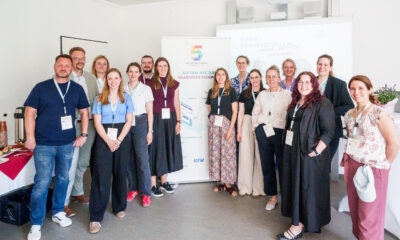 Eine Gruppe von 14 Personen steht für ein gemeinsames Foto in einem hellen Veranstaltungsraum. Die Teilnehmenden tragen Namensschilder an Schlüsselbändern. Im Hintergrund stehen ein Roll-up-Banner der Initiative „5 für Südwestfalen – Smart Cities“ und eine Präsentationsleinwand.