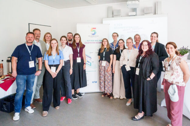 Eine Gruppe von 14 Personen steht für ein gemeinsames Foto in einem hellen Veranstaltungsraum. Die Teilnehmenden tragen Namensschilder an Schlüsselbändern. Im Hintergrund stehen ein Roll-up-Banner der Initiative „5 für Südwestfalen – Smart Cities“ und eine Präsentationsleinwand.