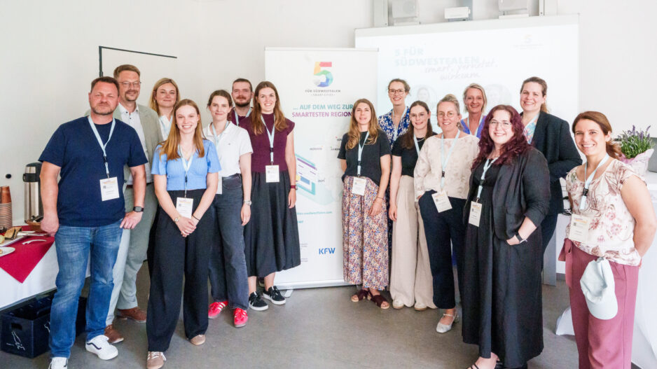 Eine Gruppe von 14 Personen steht für ein gemeinsames Foto in einem hellen Veranstaltungsraum. Die Teilnehmenden tragen Namensschilder an Schlüsselbändern. Im Hintergrund stehen ein Roll-up-Banner der Initiative „5 für Südwestfalen – Smart Cities“ und eine Präsentationsleinwand.
