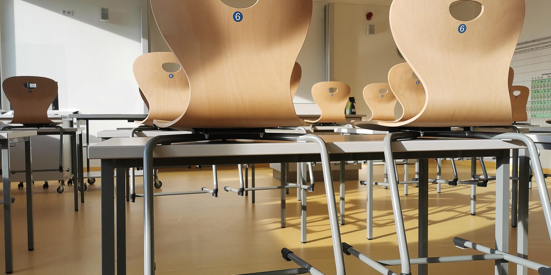 Leerer, sonnendurchfluteter Klassenraum mit mehreren Tischen. Die Holzstühle sind nach Unterrichtsende umgedreht auf die Tischplatten gestellt.