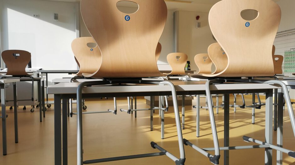 Leerer, sonnendurchfluteter Klassenraum mit mehreren Tischen. Die Holzstühle sind nach Unterrichtsende umgedreht auf die Tischplatten gestellt.