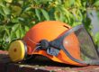 Ein orangefarbener Schutzhelm mit gelbem Gehörschutz und heruntergeklapptem schwarzem Gesichtsschutz aus Metallgitter liegt auf einer Backsteinmauer. Im Hintergrund sind grüne Blätter zu sehen.