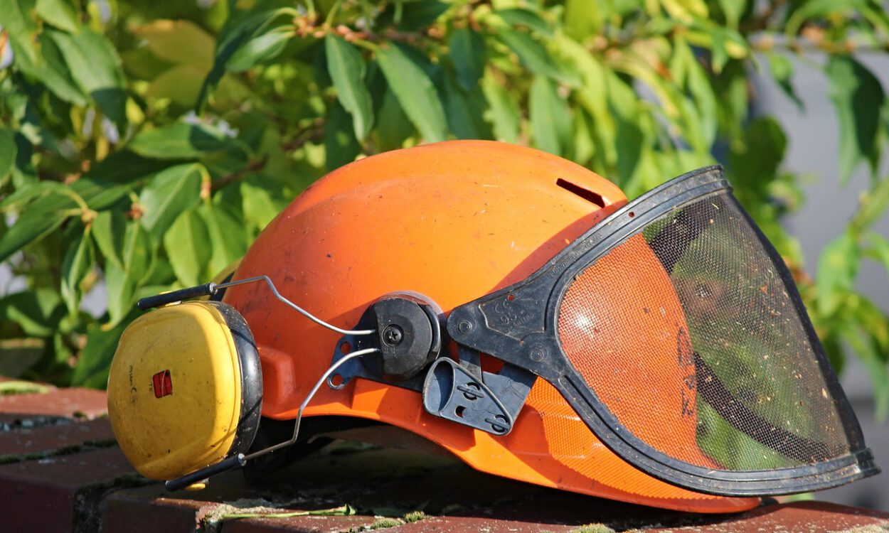 Ein orangefarbener Schutzhelm mit gelbem Gehörschutz und heruntergeklapptem schwarzem Gesichtsschutz aus Metallgitter liegt auf einer Backsteinmauer. Im Hintergrund sind grüne Blätter zu sehen.