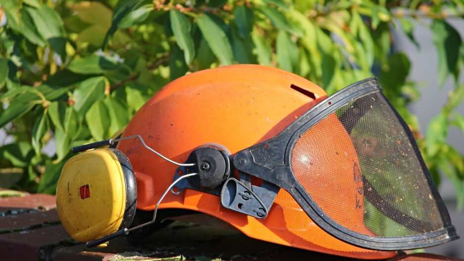Ein orangefarbener Schutzhelm mit gelbem Gehörschutz und heruntergeklapptem schwarzem Gesichtsschutz aus Metallgitter liegt auf einer Backsteinmauer. Im Hintergrund sind grüne Blätter zu sehen.