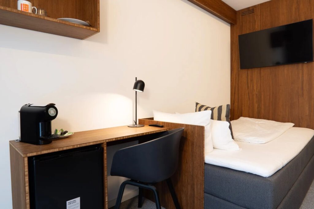 Ein modern eingerichtetes Hotelzimmer mit einem Einzelbett, weißer Bettwäsche und grauem Bettrahmen. Neben dem Bett befinden sich ein Schreibtisch aus Holz mit schwarzem Stuhl, eine Kaffeemaschine, ein kleiner Kühlschrank und eine Tischlampe. An der holzverkleideten Wand hängt ein Flachbildfernseher.